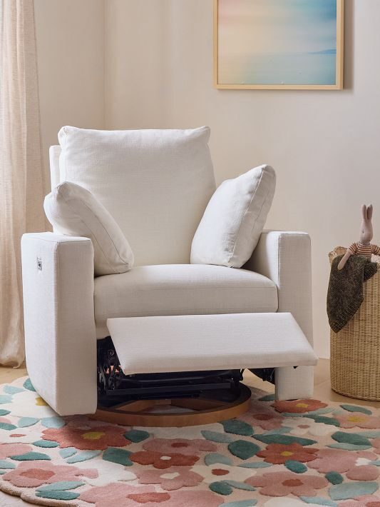 White armchair with an extended footrest sits on a floral rug. A gold floor lamp and a woven basket are nearby, with a pastel art piece on the wall. Natural light filters through curtains.