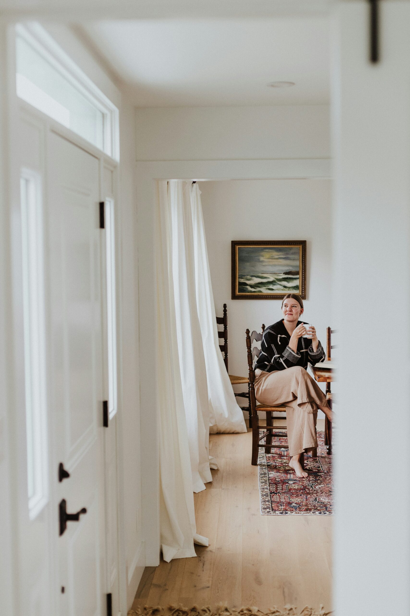 Person seated at a dining table, holding a mug. The room has wooden flooring, a colorful rug, white walls, a curtain, and a painting on the wall.