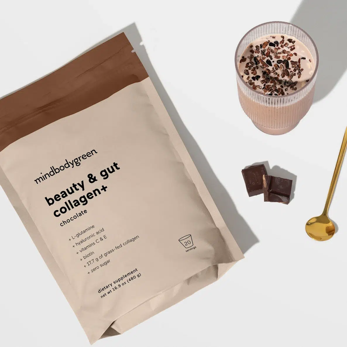 A package of mindbodygreen beauty & gut collagen+ chocolate flavor next to a glass of smoothie, two chocolate pieces, and a gold spoon.