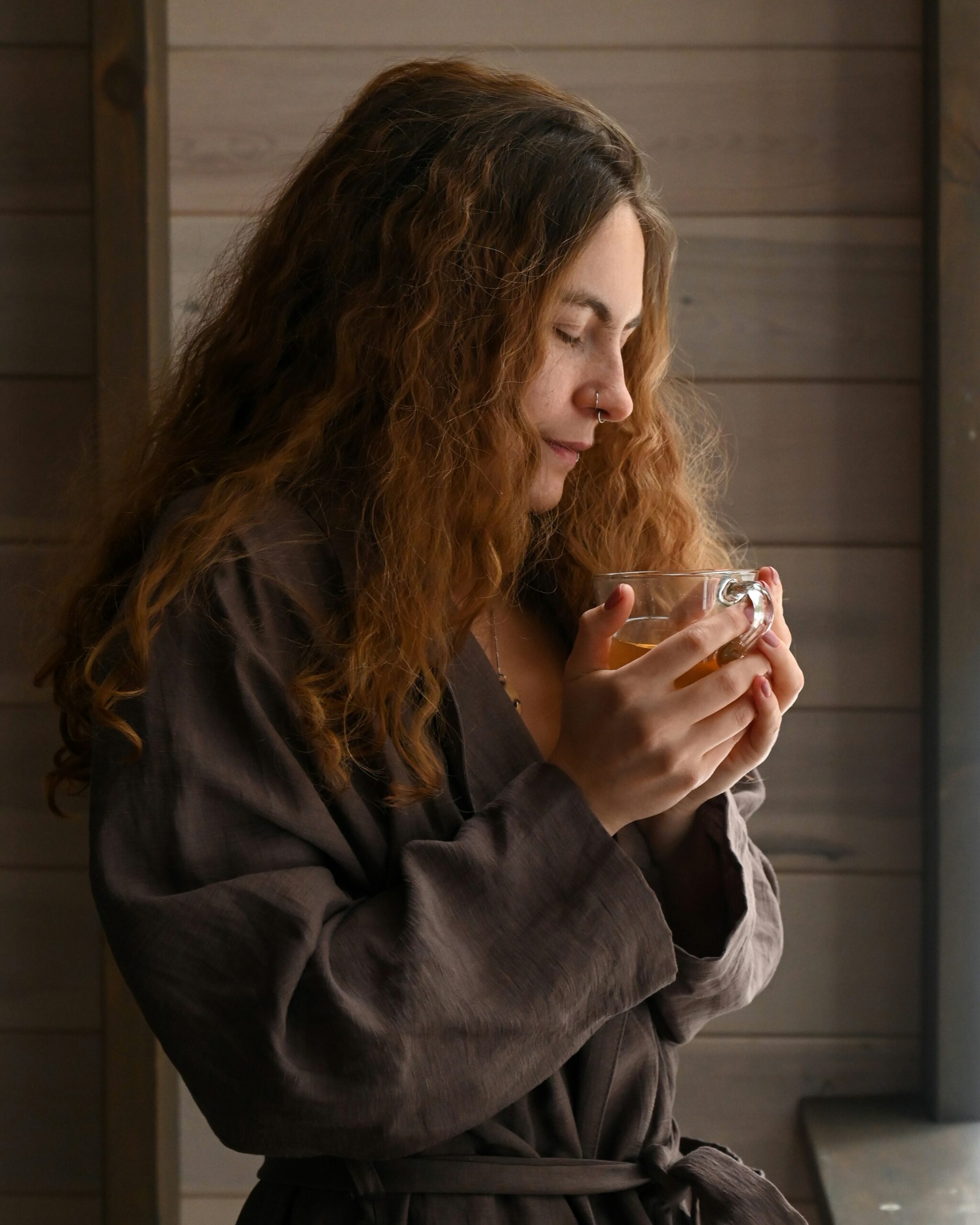 A woman with long curly hair holds a clear glass cup, standing by a window. She wears a brown robe and appears to be smelling or sipping the beverage inside.