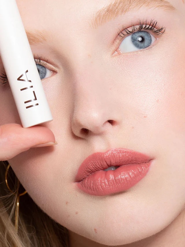 Close-up of a person holding a white ILIA product near their face. The focus is on their eyes and lips with a natural makeup look.