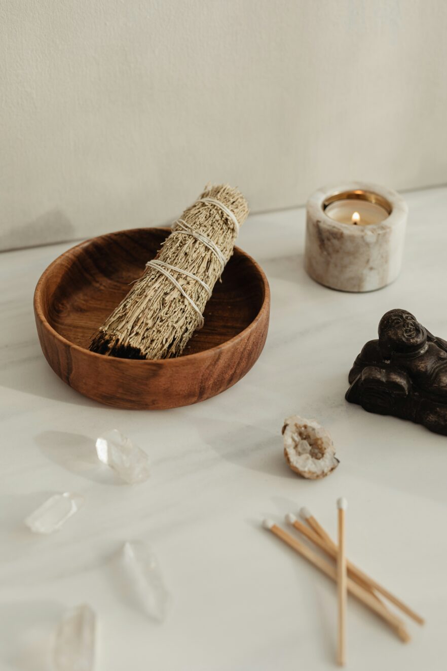 A wooden bowl holds a sage bundle on a table with a lit candle, incense sticks, crystals, a sliced geode, and a small statue.