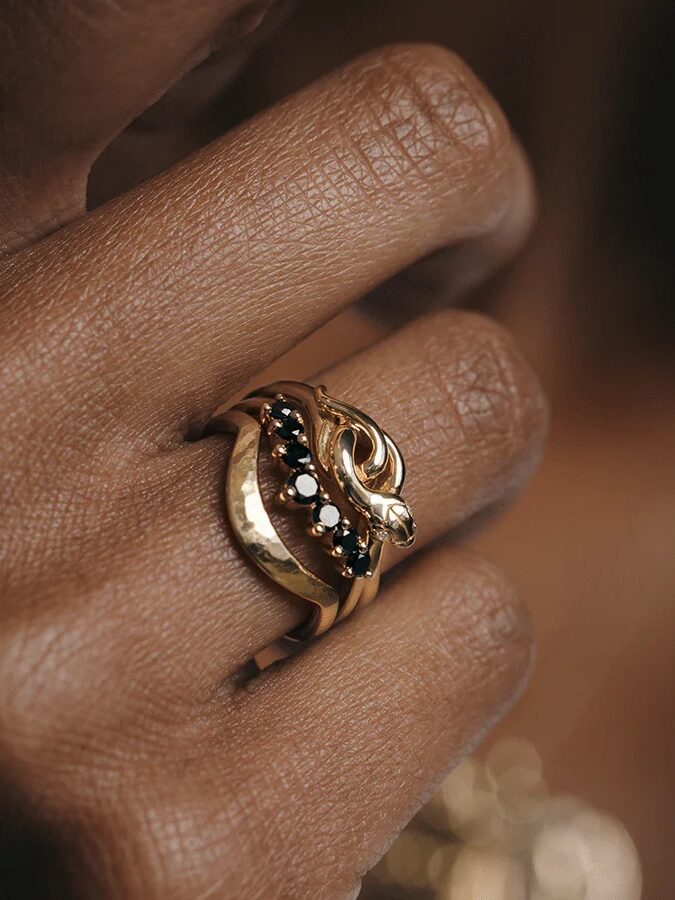 Close-up of a hand wearing a gold ring shaped like a snake, adorned with small black gemstones.