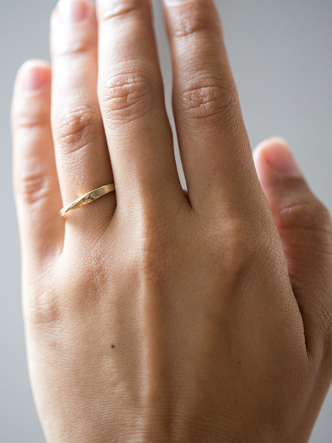 A hand wearing a simple gold ring on the ring finger, with a neutral background.