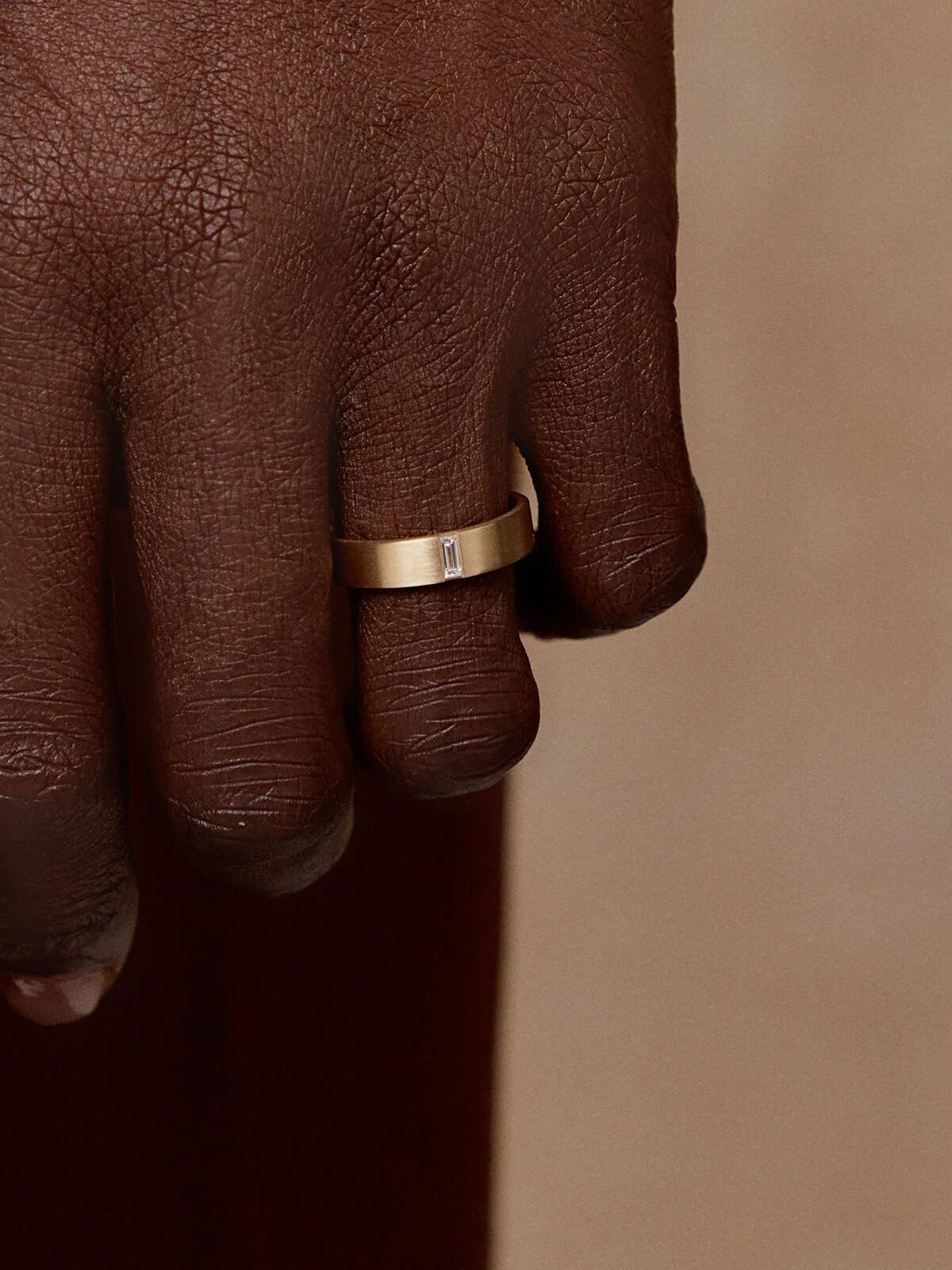 A hand wearing a simple gold band ring with a small rectangular stone, against a brown background.