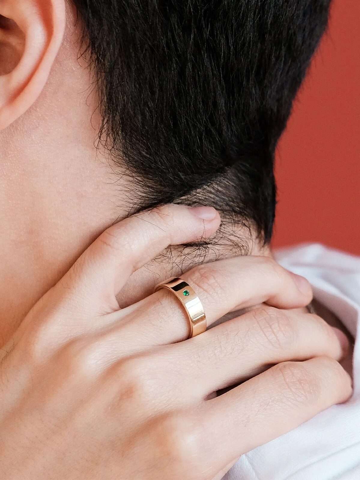 Close-up of a person touching the back of their neck. The person is wearing a gold ring on their hand. The background is solid red.