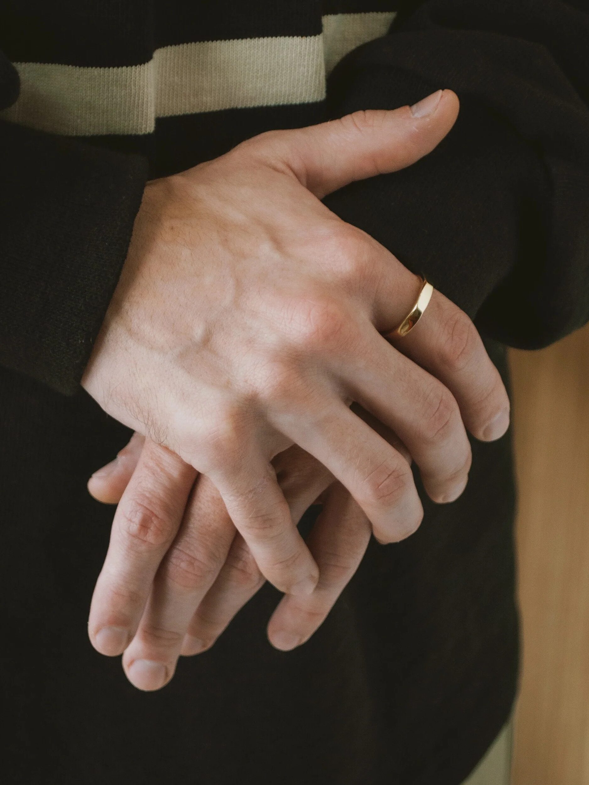 A person wearing a black sweater rests their hands, one with a gold ring, in front of them.