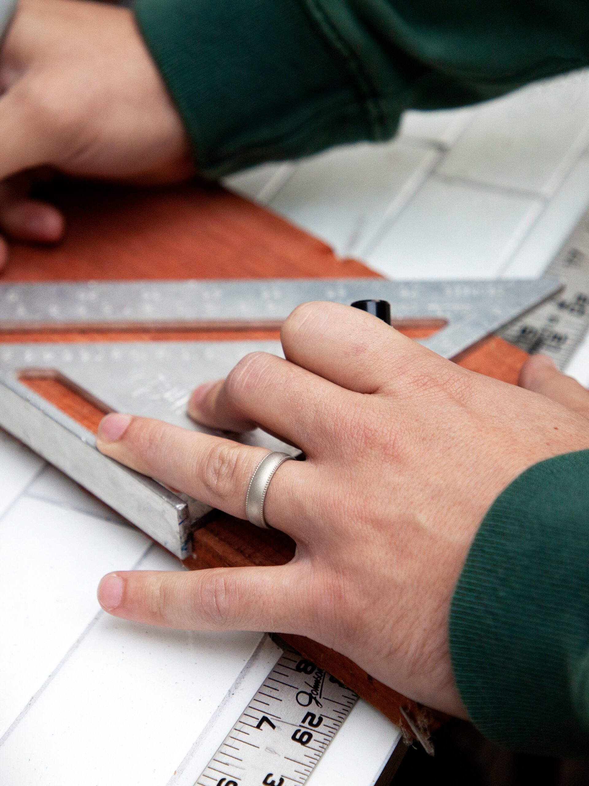 Hands using a metal square ruler and pen on a wooden surface, with a multi-tool and measuring tape nearby.