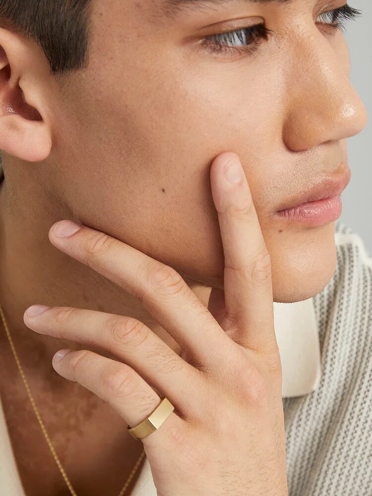 A person with short hair touches their face with their hand, wearing a gold ring and necklace, while gazing sideways.