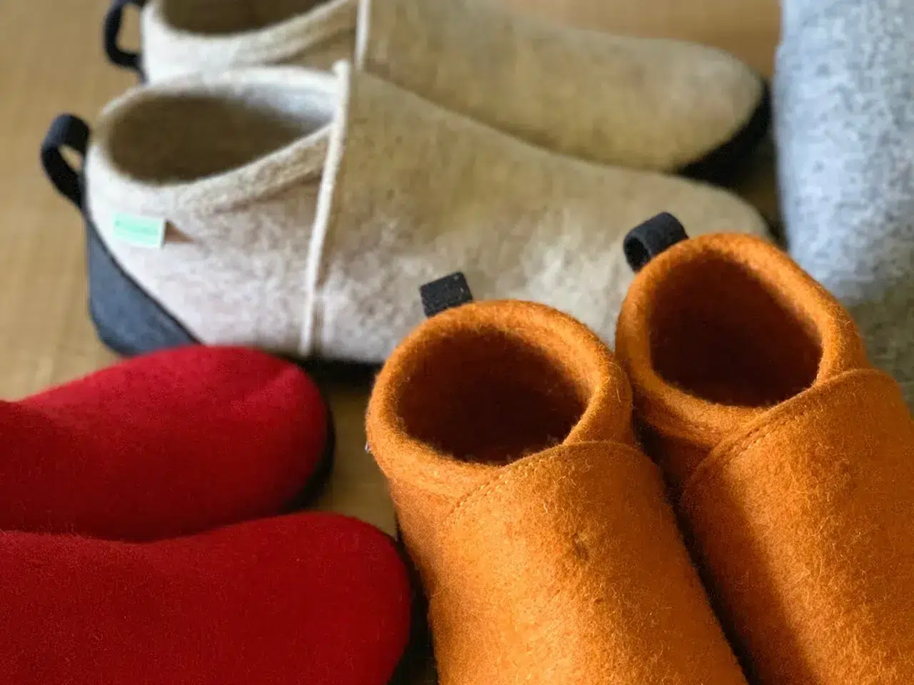 Various colorful felt slippers are displayed on a wooden surface, including pairs in orange, red, beige, and gray.