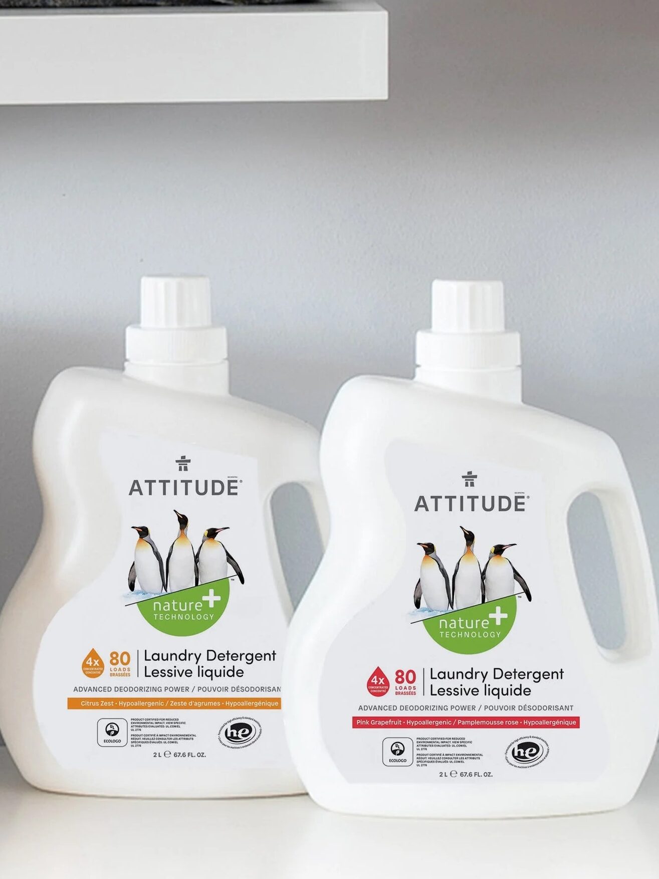 Two white bottles of Attitude laundry detergent are placed on a counter. Labels indicate scents: Wildflowers, Pink Grapefruit, and Citrus Zest. A woven basket sits beside the bottles.