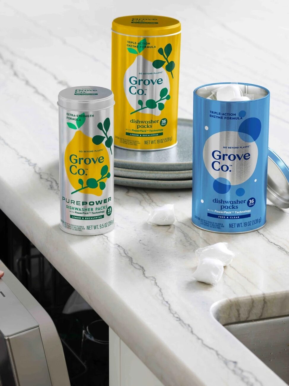 A hand opens a dishwasher next to three Grove Co. cleaning product containers on a kitchen counter, with a few dishwasher packs visible nearby.