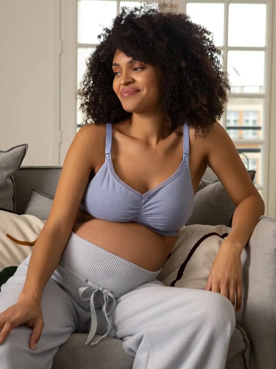 Pregnant woman with curly hair sits on a couch in a softly lit room, wearing a light blue top and gray pants, looking to the side with a gentle smile.