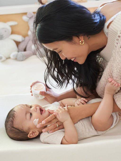 A woman applies cream to a baby lying on a bed in a cozy room with stuffed animals.