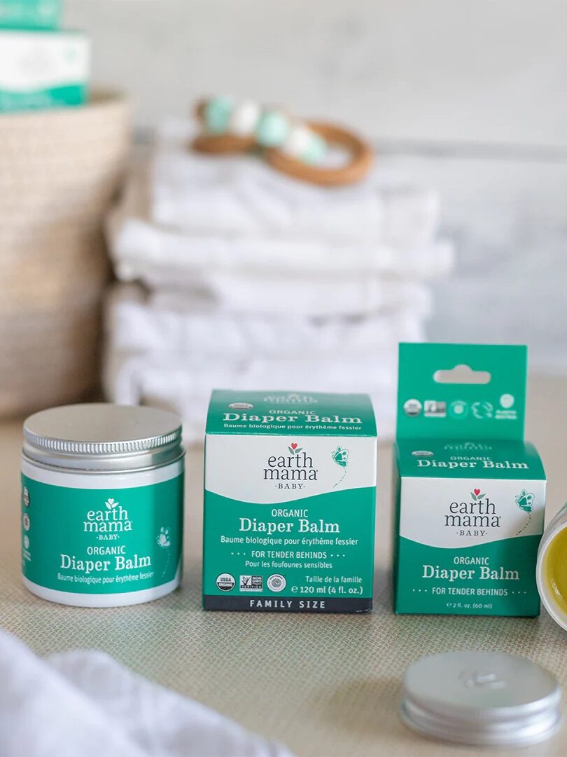 The image shows three containers of Earth Mama Organics diaper balm on a surface. One is open, displaying a yellow balm. Stacked diapers are blurred in the background.