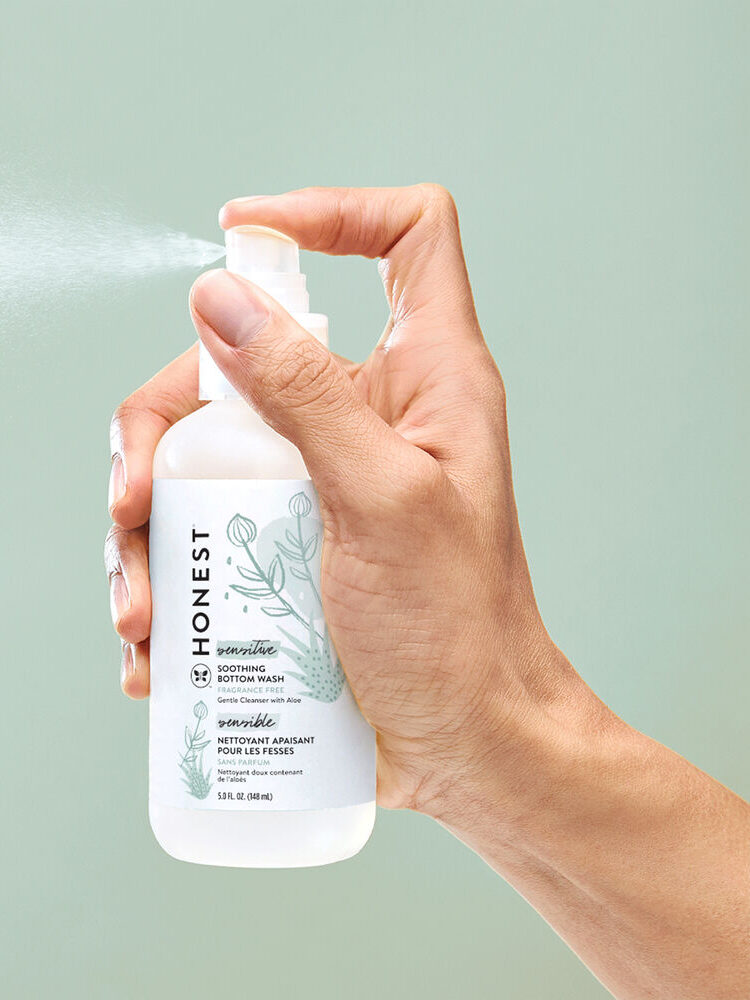 A hand sprays a bottle of Honest brand soothing therapy lotion against a light green background.