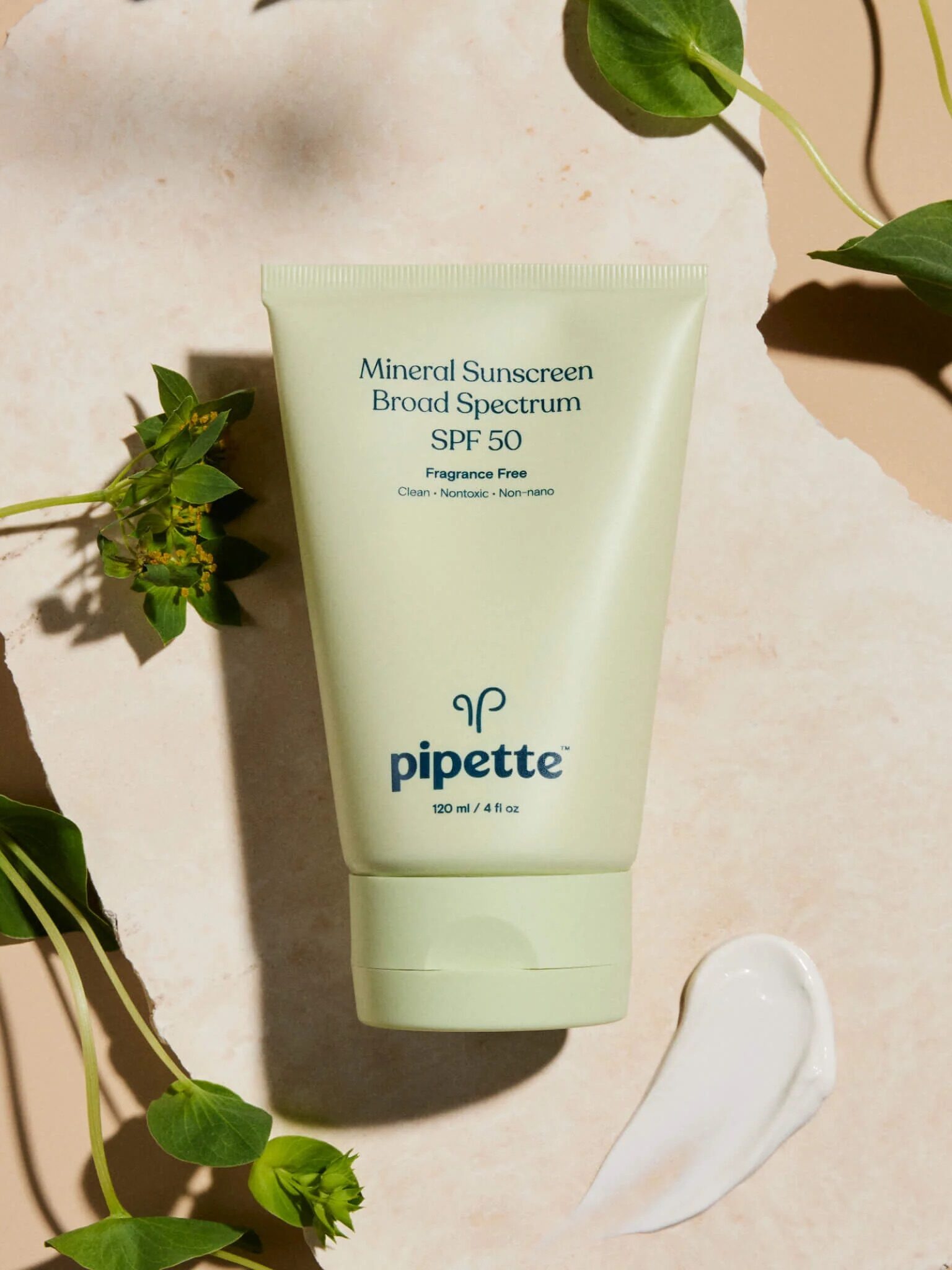 A tube of Pipette Mineral Sunscreen Broad Spectrum SPF 50 on a beige background with green leaves and a dollop of sunscreen next to it.