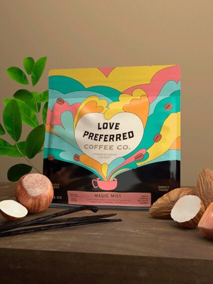 A colorful coffee package labeled "Love Preferred Coffee Co." is surrounded by vanilla pods, hazelnuts, almonds, caramel pieces, and leaves on a wooden surface.