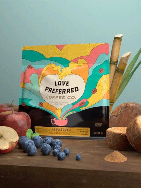 A colorful coffee bag labeled "Love Preferred Coffee Co." is surrounded by apples, blueberries, sliced sweet potatoes, sugar cane, and a green leaf on a wooden surface.