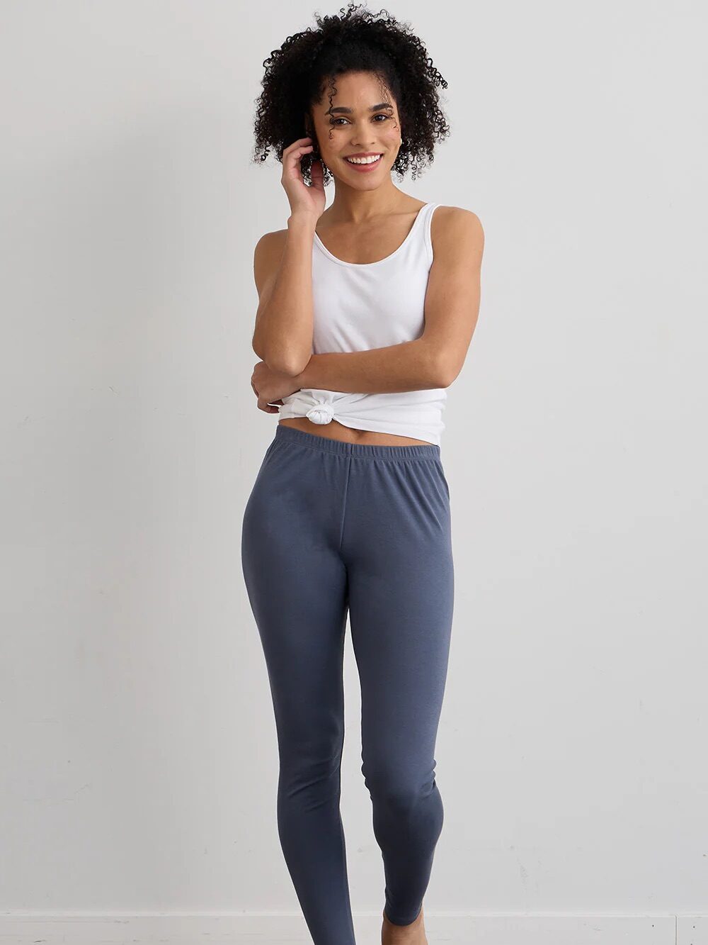 A person in a white tank top and blue leggings stands barefoot against a plain background, smiling.