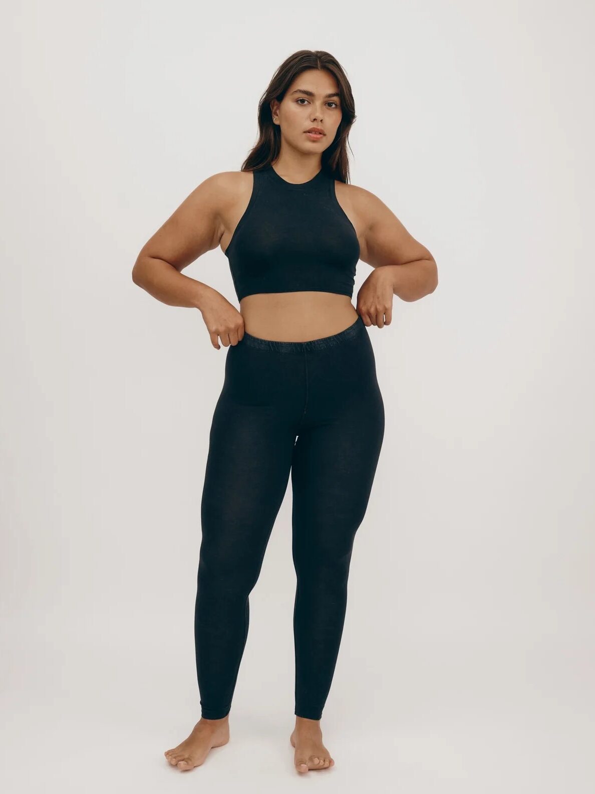 A woman stands barefoot against a plain background, wearing a black sports bra and leggings, with her hands resting at her waist.