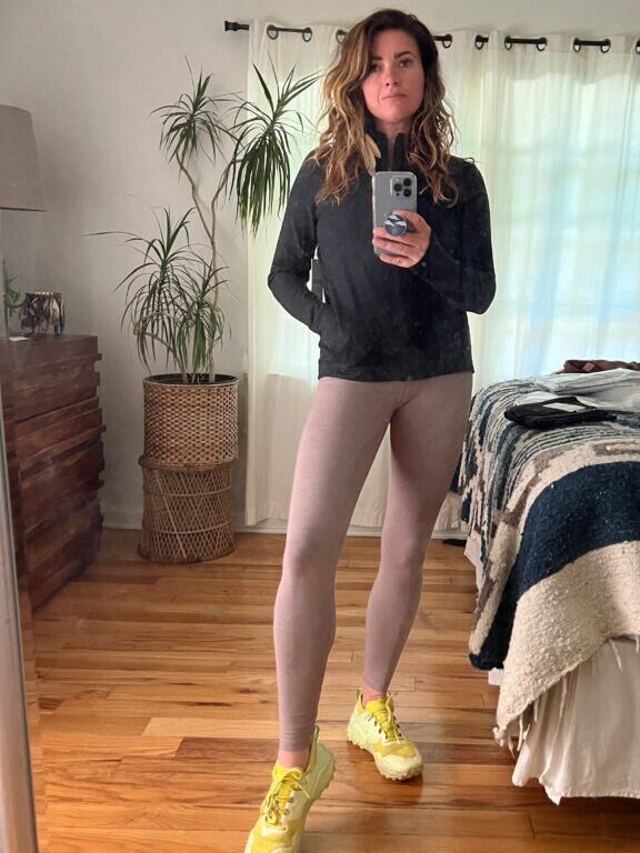 A woman stands in a bedroom wearing a black sweater, beige leggings, and yellow sneakers, holding a smartphone and posing in front of a mirror.