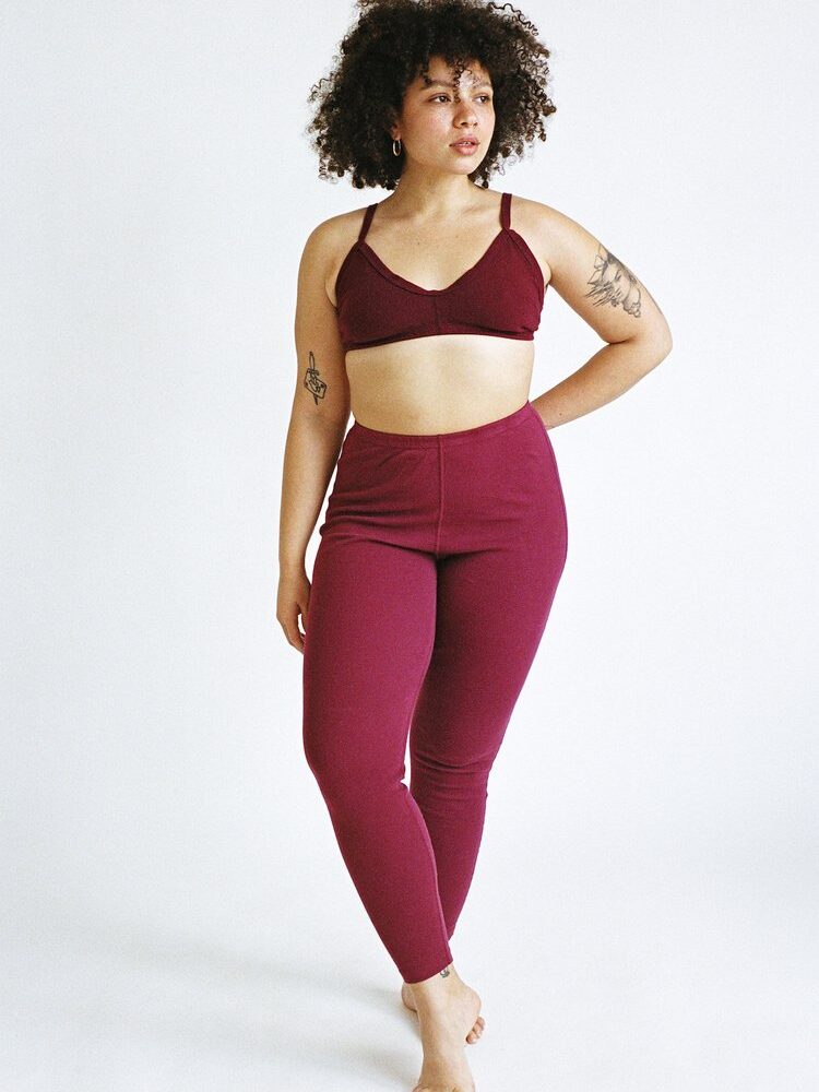 Person with curly hair wearing a maroon sports bra and leggings stands barefoot against a plain background.