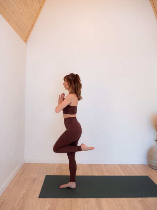 A person in brown workout attire practices a balancing yoga pose on one leg, standing on a yoga mat in a minimalistic room with light wood floors and ceiling.