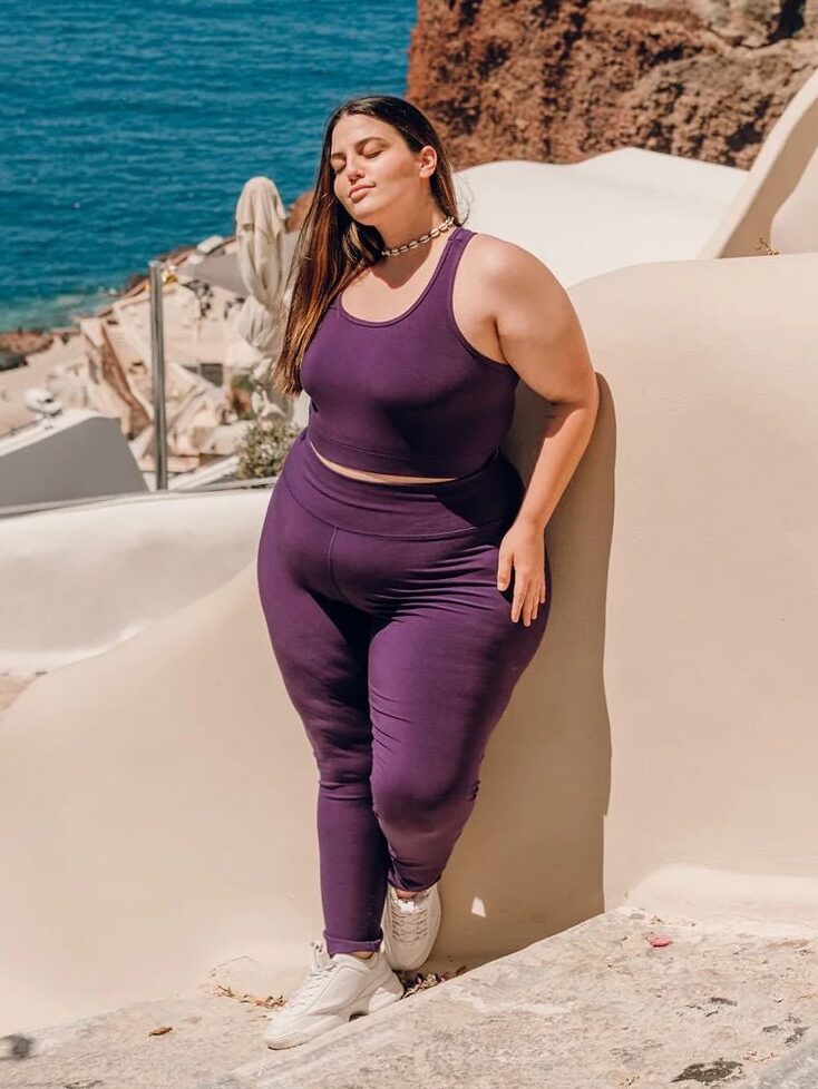 A woman in a purple outfit stands against a white wall by a cliffside, with the ocean in the background.