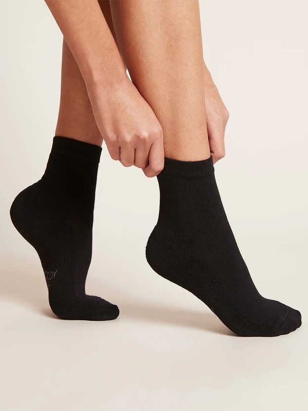 A person pulls on black organic ankle socks, showing only their lower legs and feet.