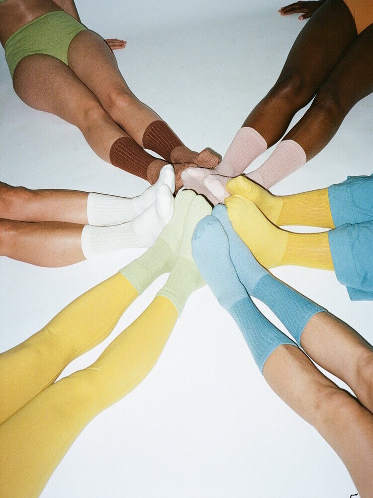 A circle of eight people sit on the floor with their legs extended and feet touching. They wear colorful organic socks and bottoms in shades of green, yellow, blue, brown, and pink.