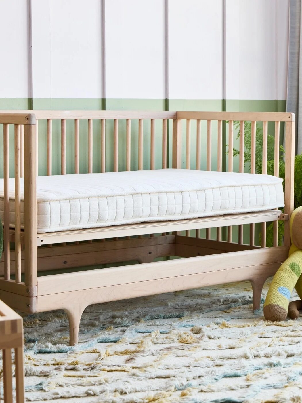 Wooden crib with a mattress next to a stuffed toy dog, on a textured rug with some plants in the background.