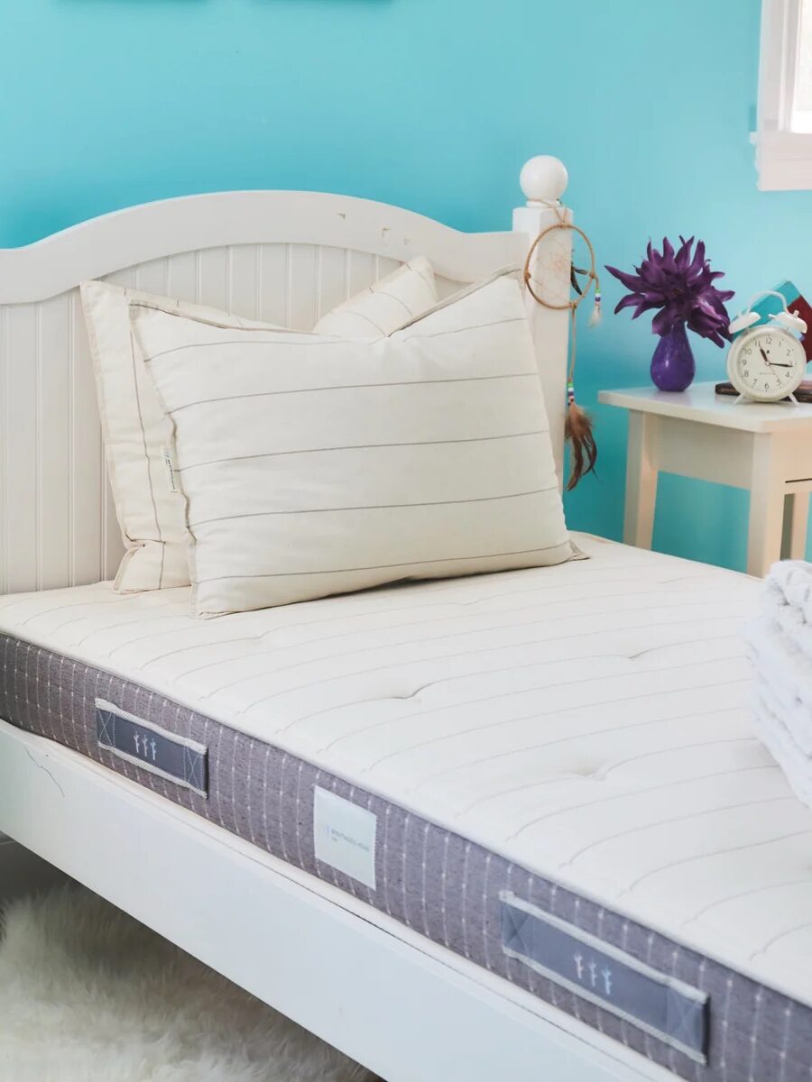 White twin bed with a beige-striped mattress and two matching pillows in a room with turquoise walls. A nightstand with a clock and a purple plant sits nearby on a wooden floor with a white rug.