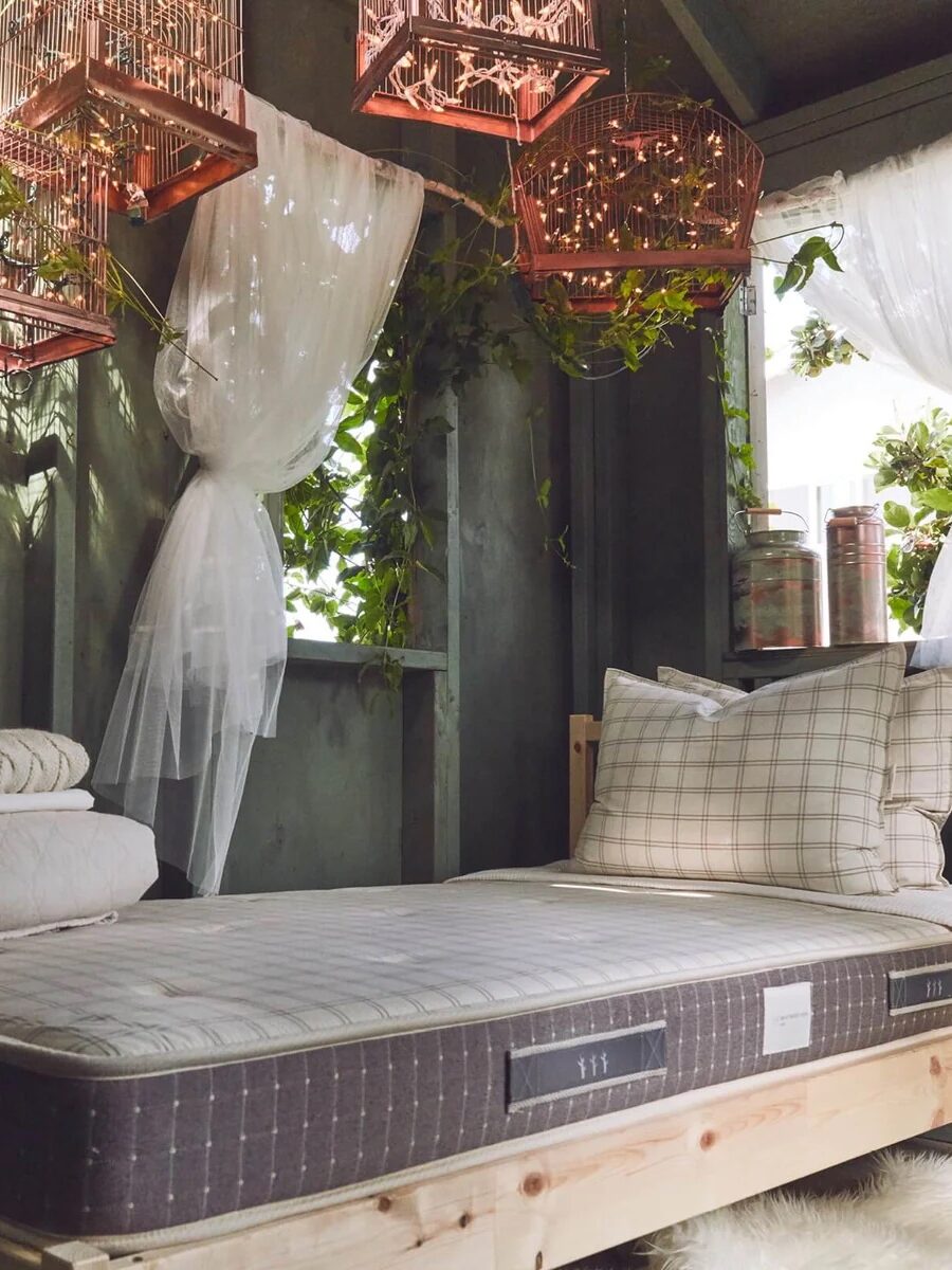 Cozy room with a neatly made bed, surrounded by greenery and hanging woven lights. Sheer curtains allow natural light to filter in.