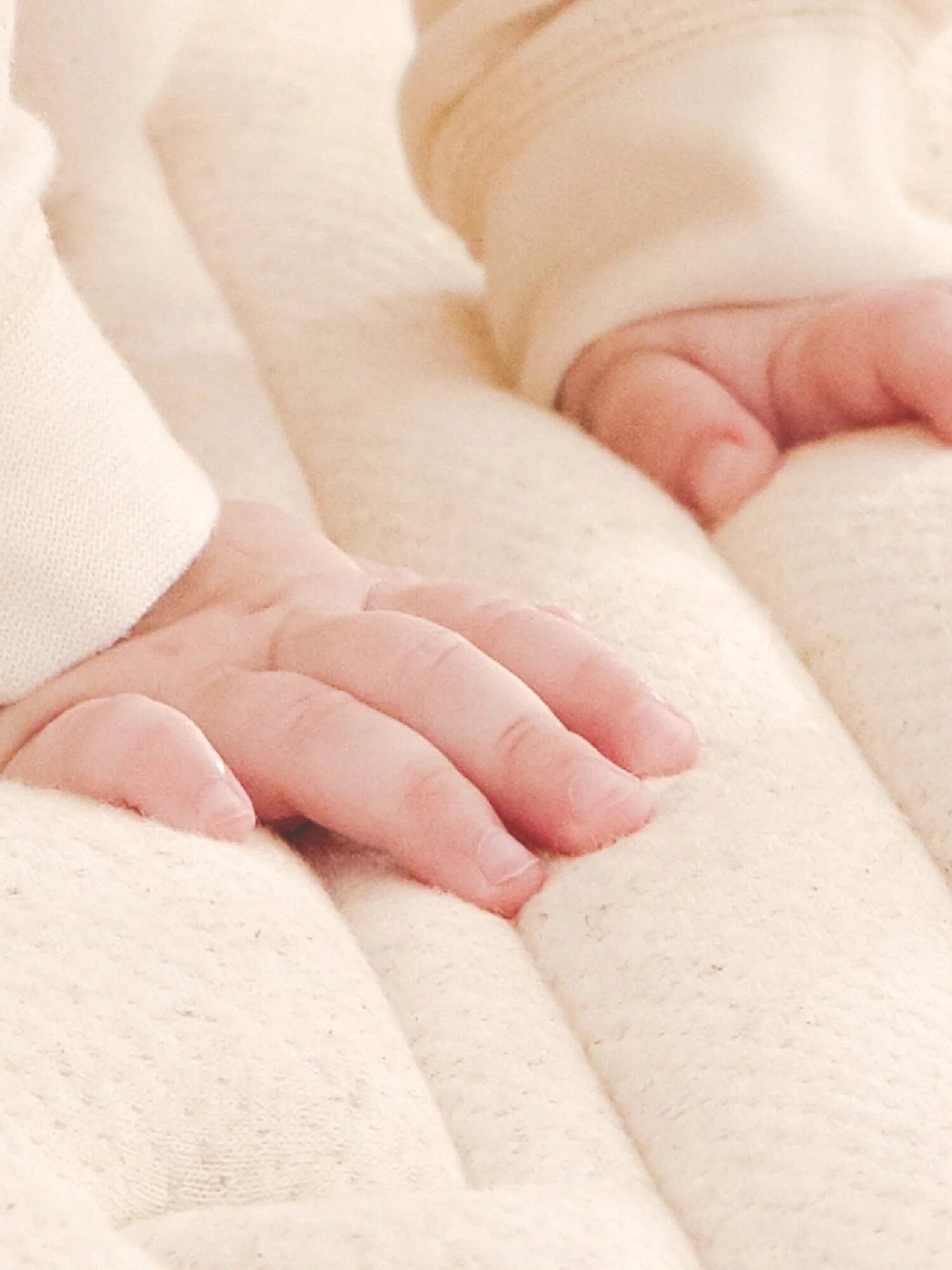 Close-up of a baby's hands pressing on a soft, cushioned surface. The baby is wearing a cream-colored outfit.