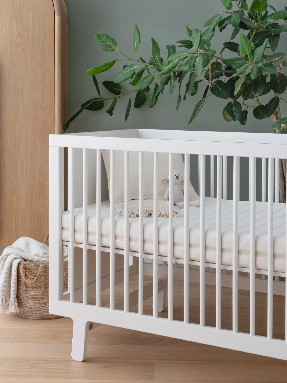 A white crib in a nursery with a green wall, wooden wardrobe, and a potted plant. A woven basket sits next to the crib with a folded blanket inside.