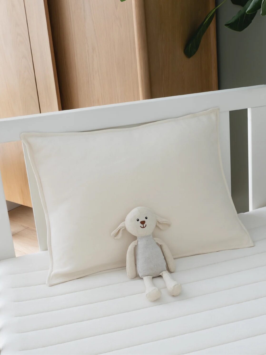 A white crib with a light-colored pillow and a small stuffed animal on the mattress. Green leaves hang from the top right corner.