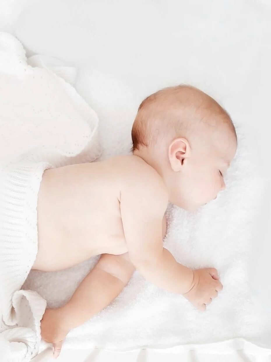 A baby sleeps on a white blanket, lying on its stomach with arms bent by its side.