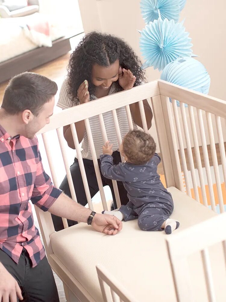 Adults playing with a baby in a crib; one adult is sitting, the other standing, with blue decorations in the background.