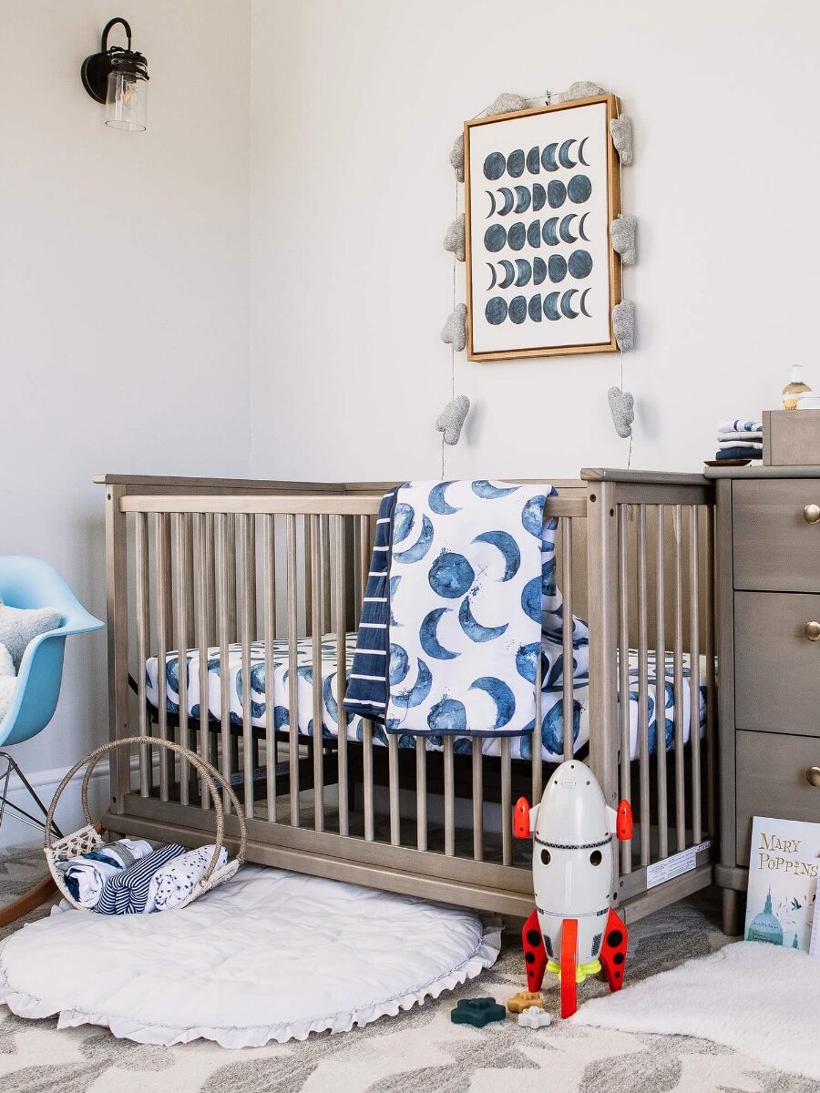 A nursery with a crib, moon-themed bedding, rocket toy, rocking chair, and dresser. A moon phase art piece hangs above the crib, and books are on the floor.