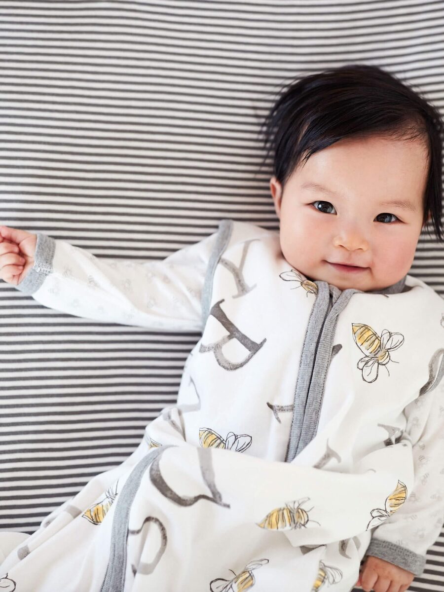 A baby in a white and gray outfit with bee patterns lies on a striped blanket, smiling slightly at the camera.