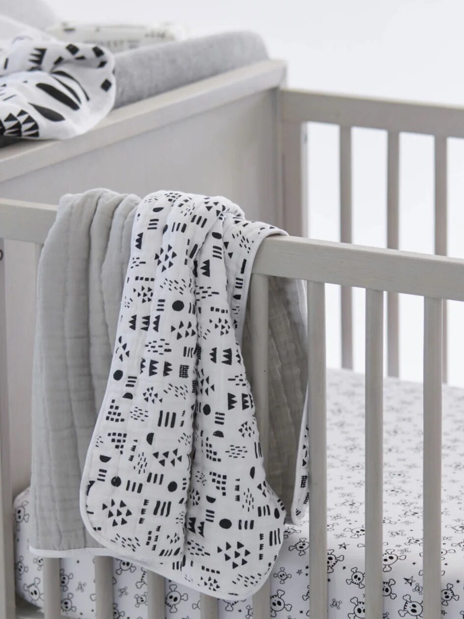 White baby crib with a patterned blanket draped over the side and a patterned mattress inside.