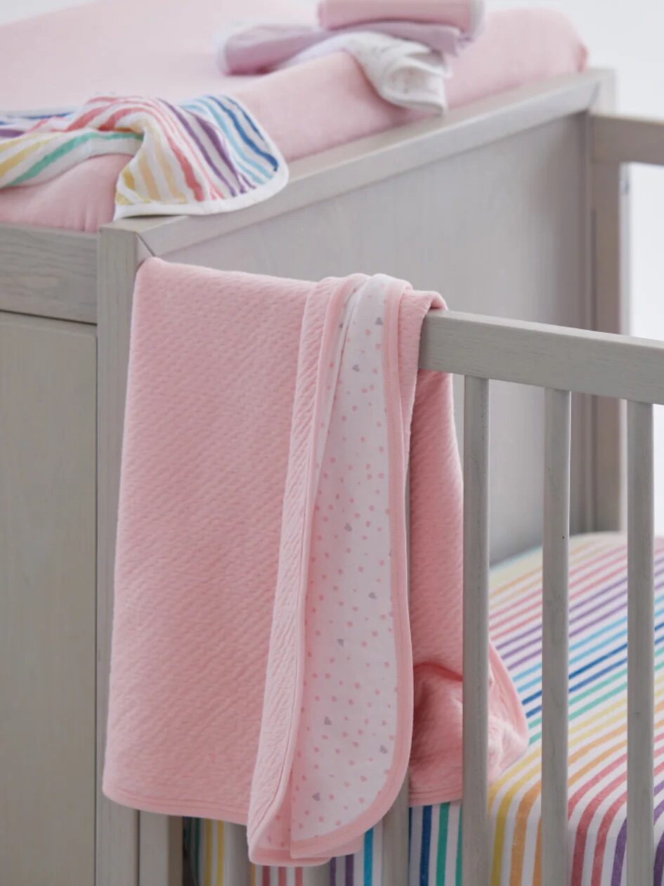 A baby crib with a pink blanket draped over the side and a rainbow-striped mattress. A matching pink cloth is on a nearby changing table.