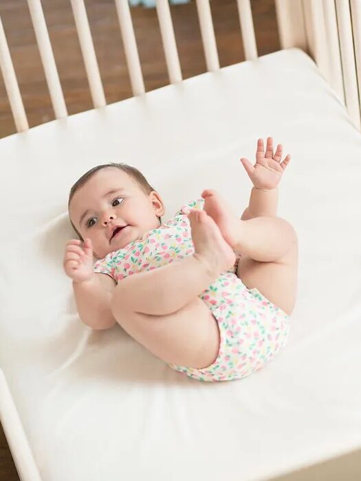 A baby in a patterned onesie lies on a white mattress in a crib, looking upward with arms raised and legs bent.