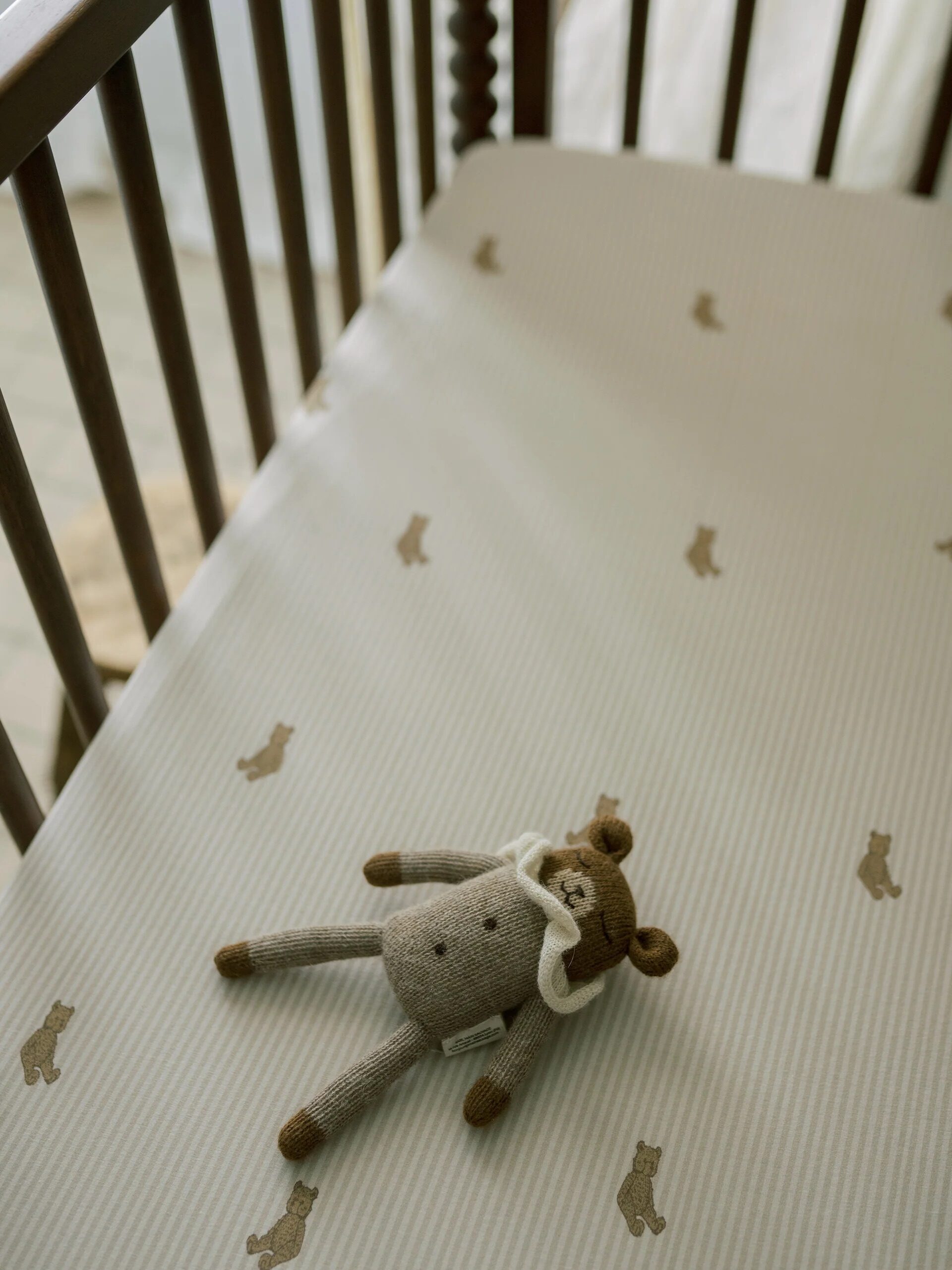 A stuffed bear toy lies on a light-colored crib sheet patterned with small teddy bears. The sheet covers a wooden crib.