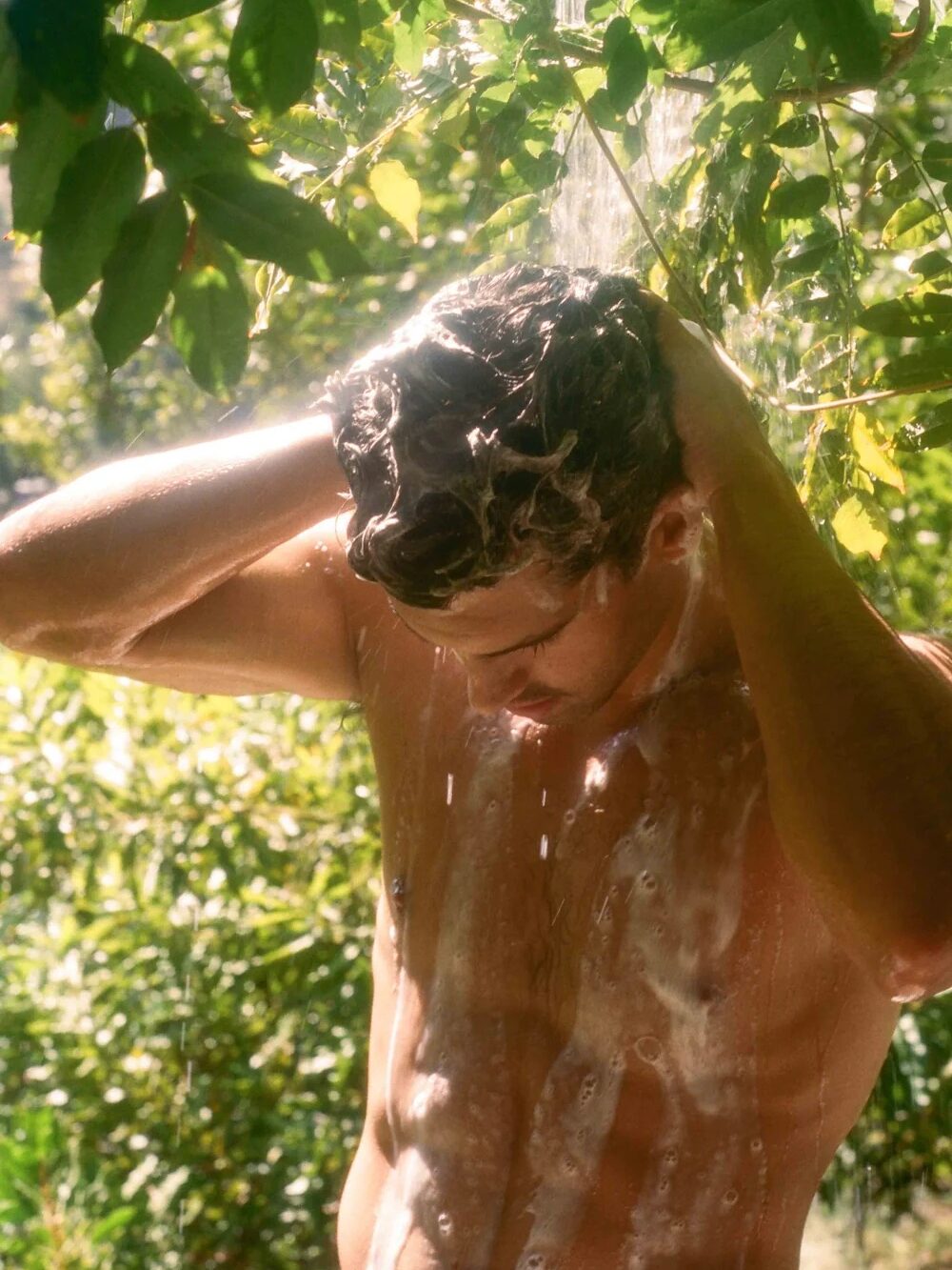 Person showering outdoors under green foliage, with water cascading down their head and soap lather on their body.