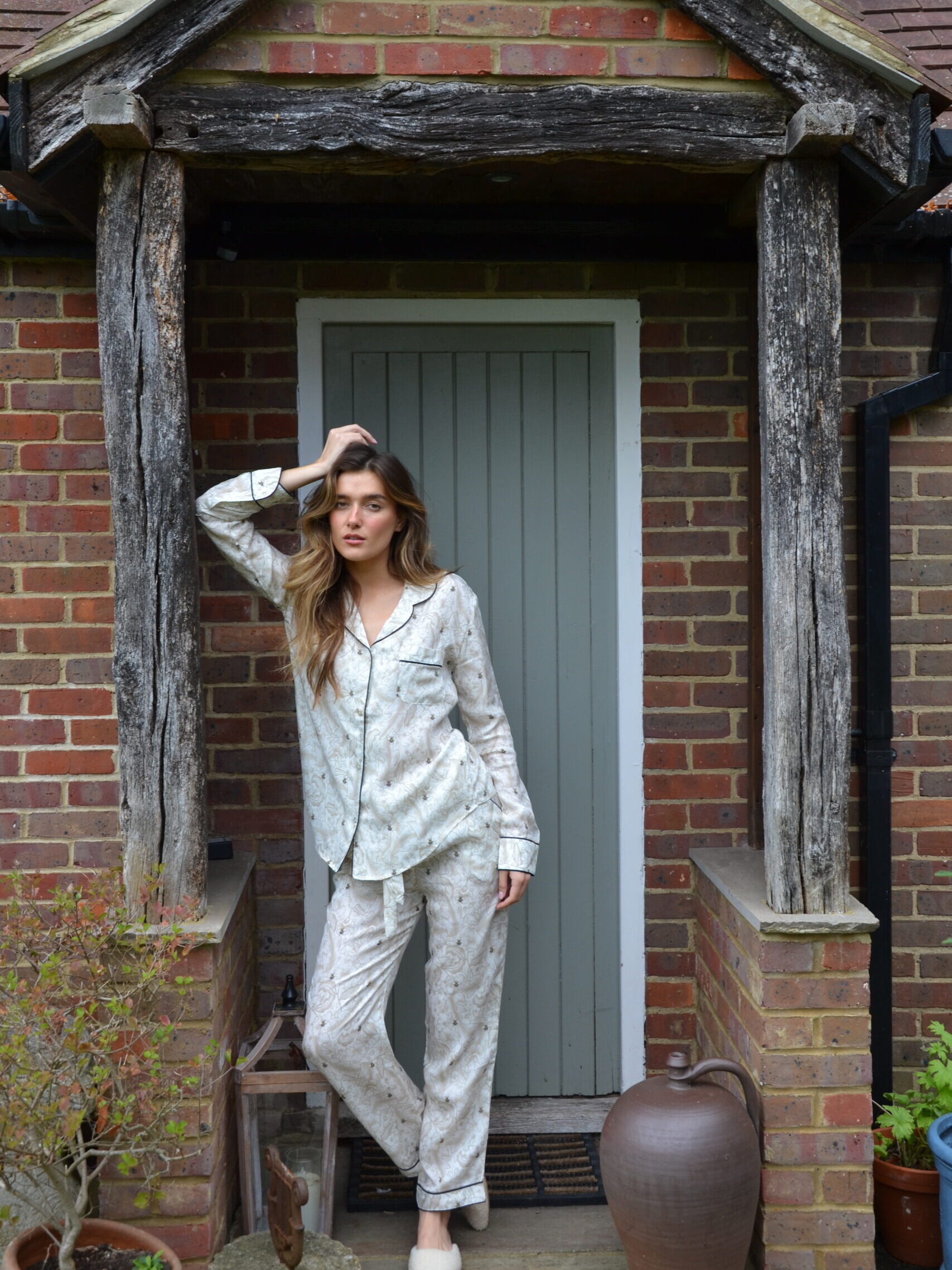 Person in patterned pajamas standing in a rustic doorway with one hand on their head. Brick wall and large clay pot nearby.