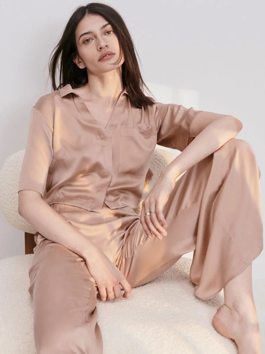 A person wearing a matching silk pajama set sits on a soft chair against a plain background.