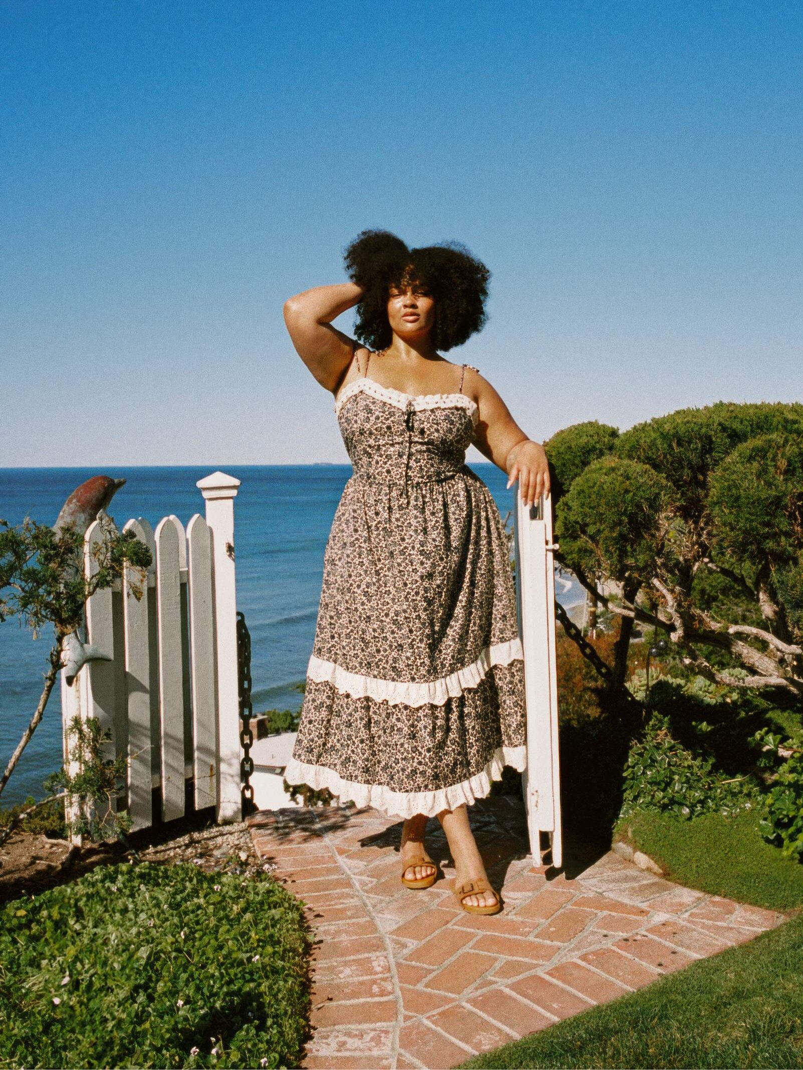 Person in a floral dress stands by a white fence with the ocean in the background.