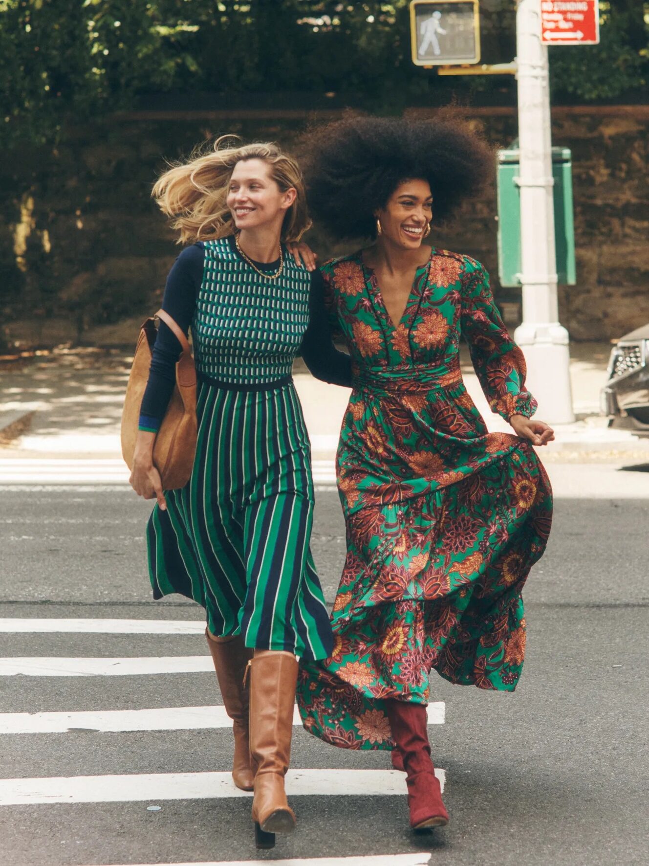 Two women in patterned dresses and boots joyfully cross a city street.